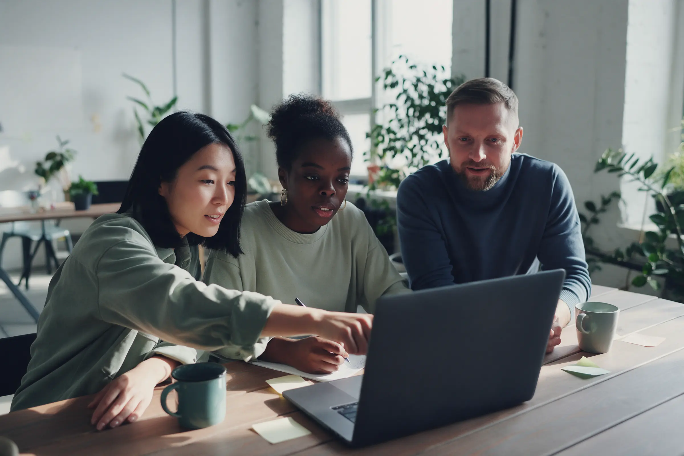 Three colleagues lean over one laptop at a long table in a bright office, one pointing at the screen