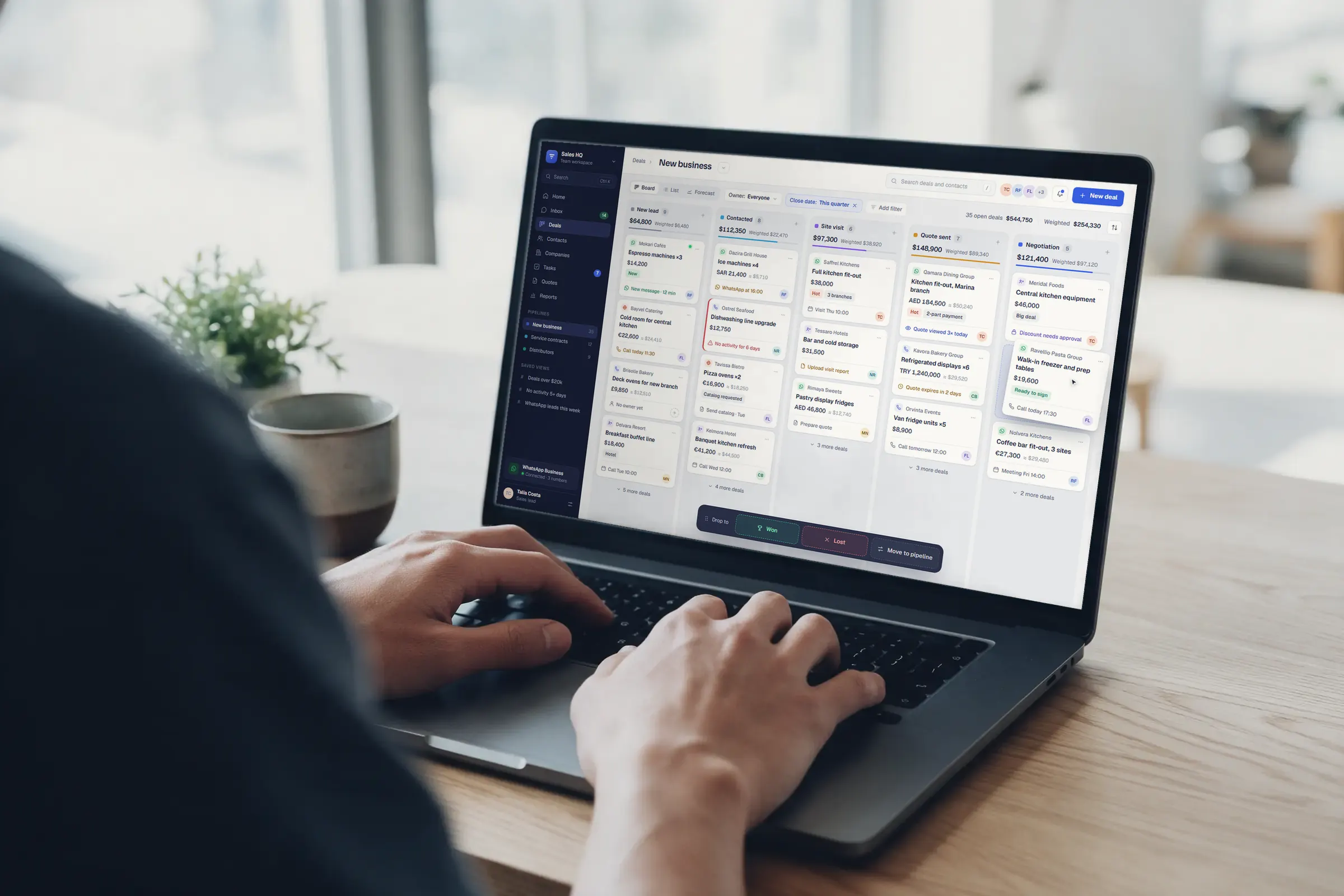 Hands typing on a laptop on a light oak desk; the screen shows the deal board of our sales CRM