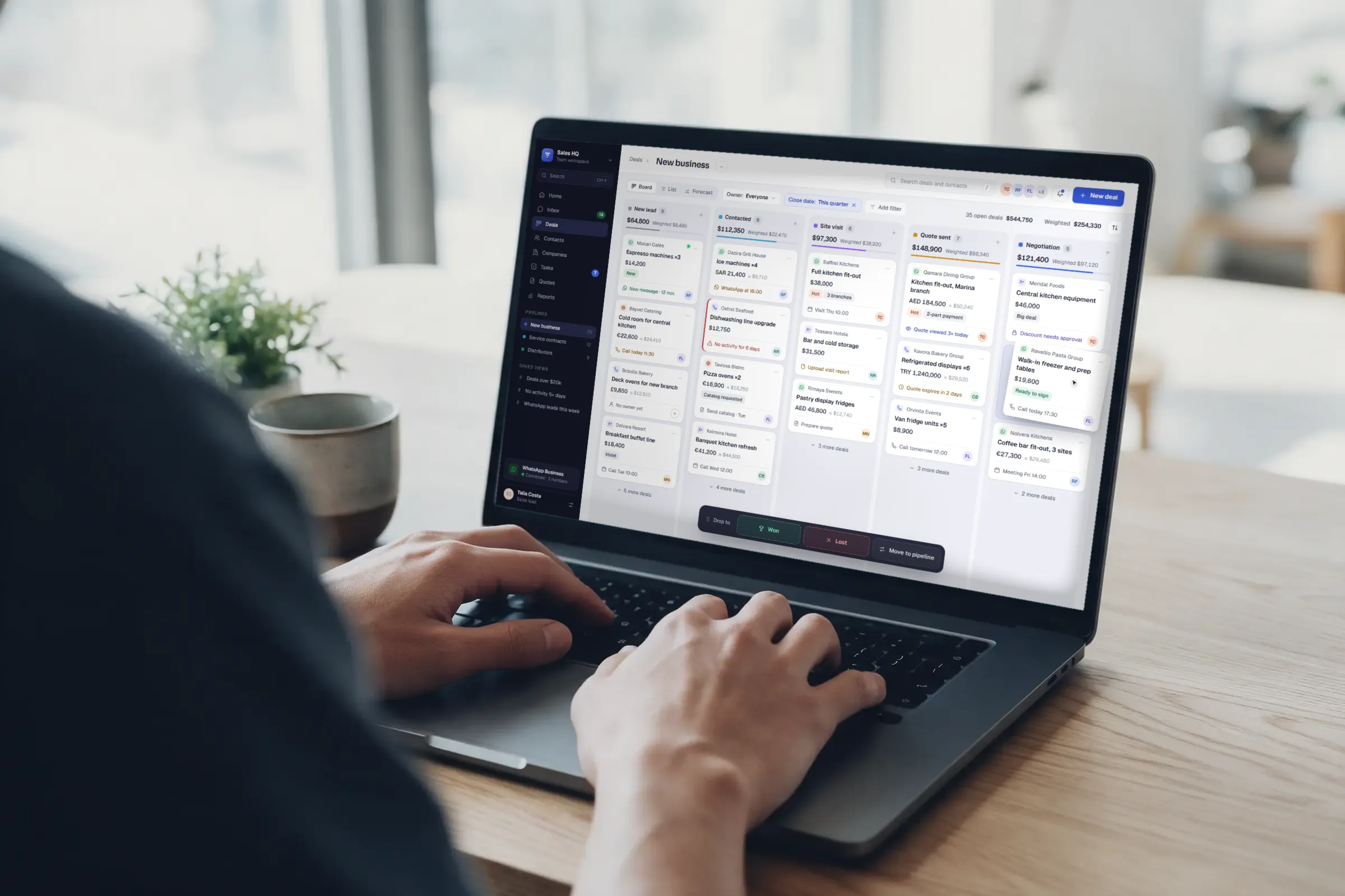 Hands typing on a laptop on a light oak desk by a bright window; the screen shows the sales CRM's deal board