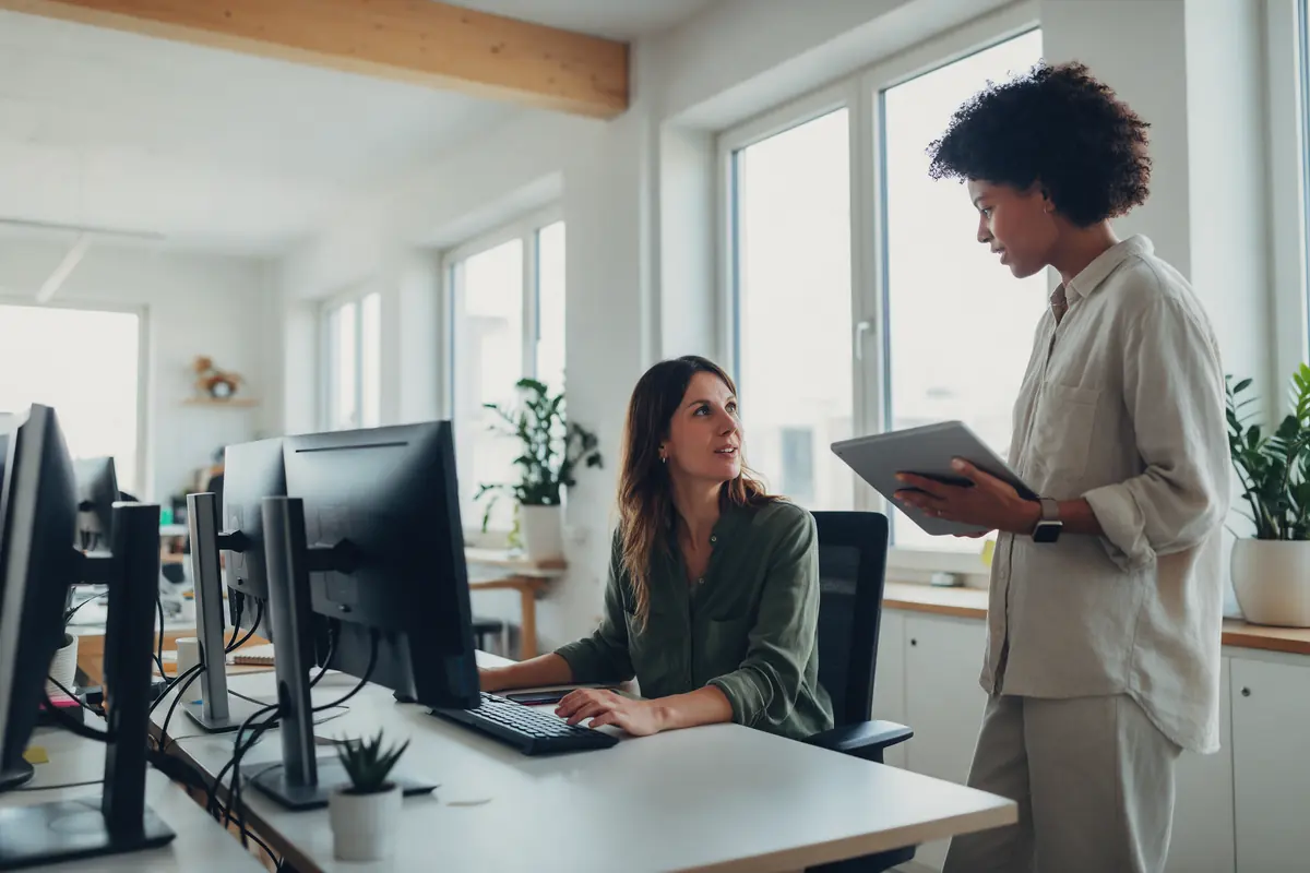 In a bright distribution office, a colleague with a tablet stops by a woman's desk to check something with her