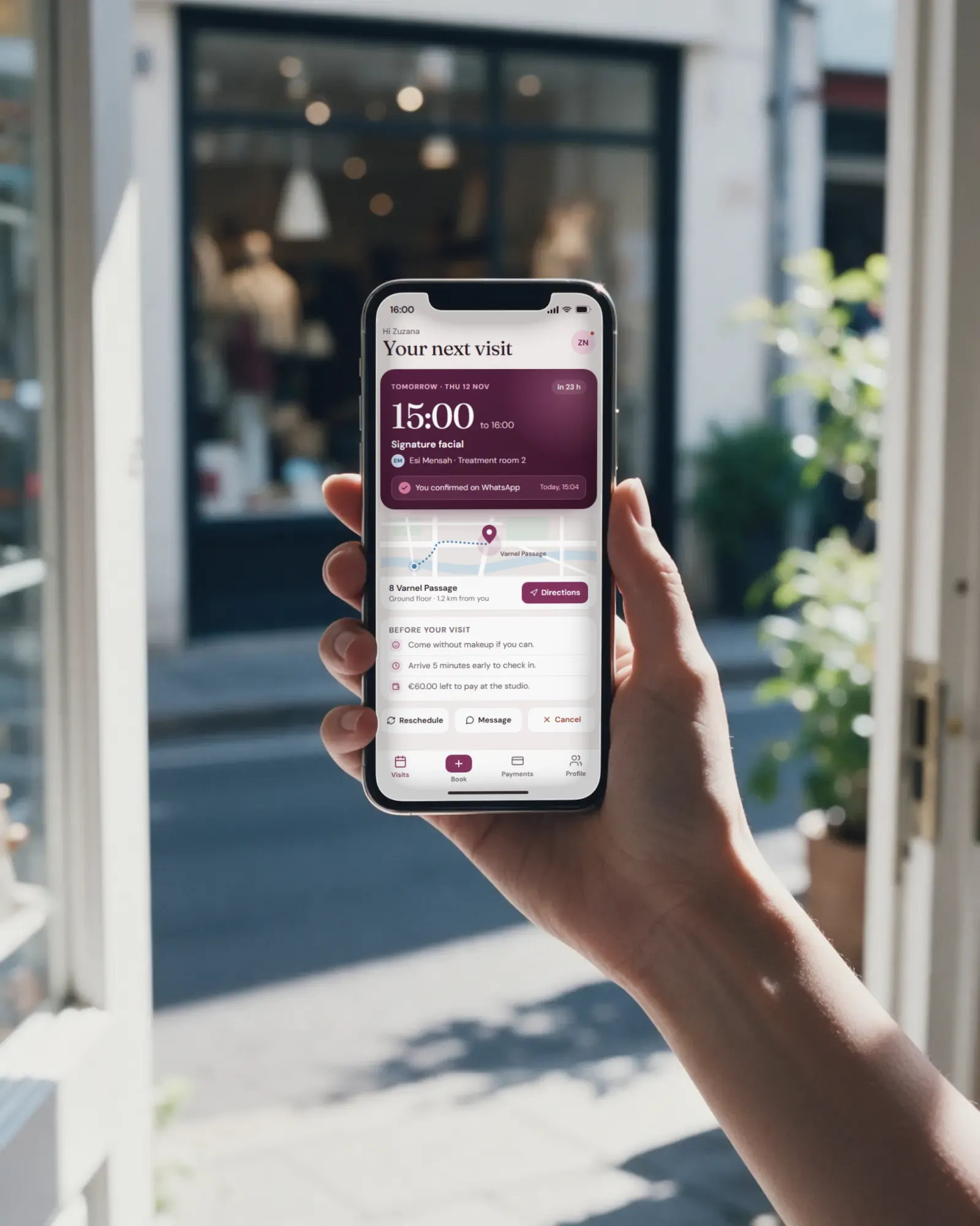 A hand holds up a phone in a bright shop doorway, showing the booking app: the client's next visit tomorrow at 15:00, confirmed on WhatsApp